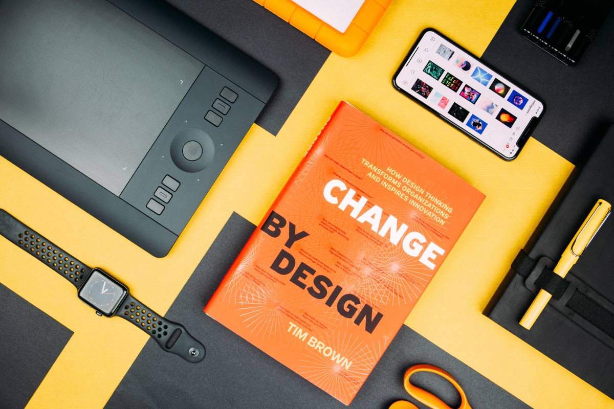 Aerial shot of a book called "Change by Design, by Tim Brown" with other tech such as phone, smart watch, and tablet surrounding it.
