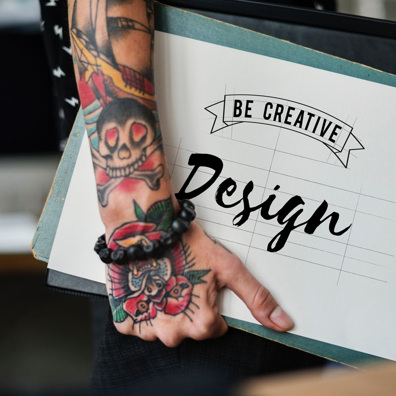 Person with tattoo arm sleeve holding a paper graphic that says, "Be Creative" on ribbon banner, and "Design" under that text.